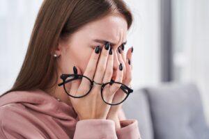 Woman with black nails and pink hoodie, with her glasses in her hand, rubbing her dry and tired eyes